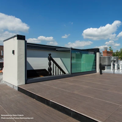 A spacious rooftop terrace featuring wooden flooring, glass railings, and a staircase leading to an interior space. The sky is partly cloudy, enhancing the bright and open atmosphere.
