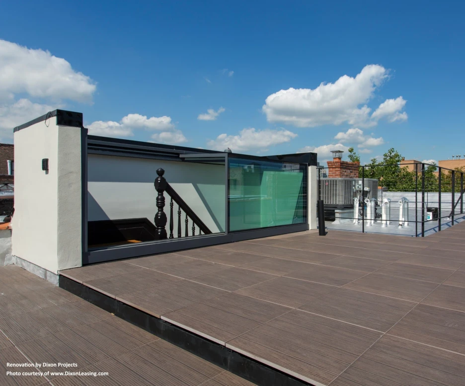 A rooftop space featuring wooden decking, glass railings, and a staircase. Bright blue sky with a few fluffy clouds is visible in the background.