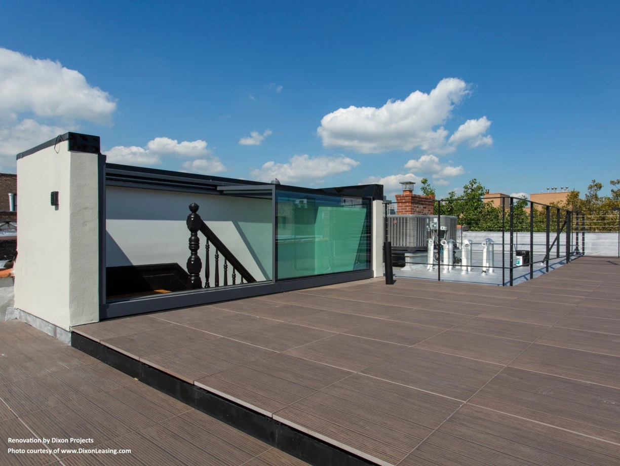 A rooftop space featuring wooden decking, glass railings, and a staircase. Bright blue sky with a few fluffy clouds is visible in the background.