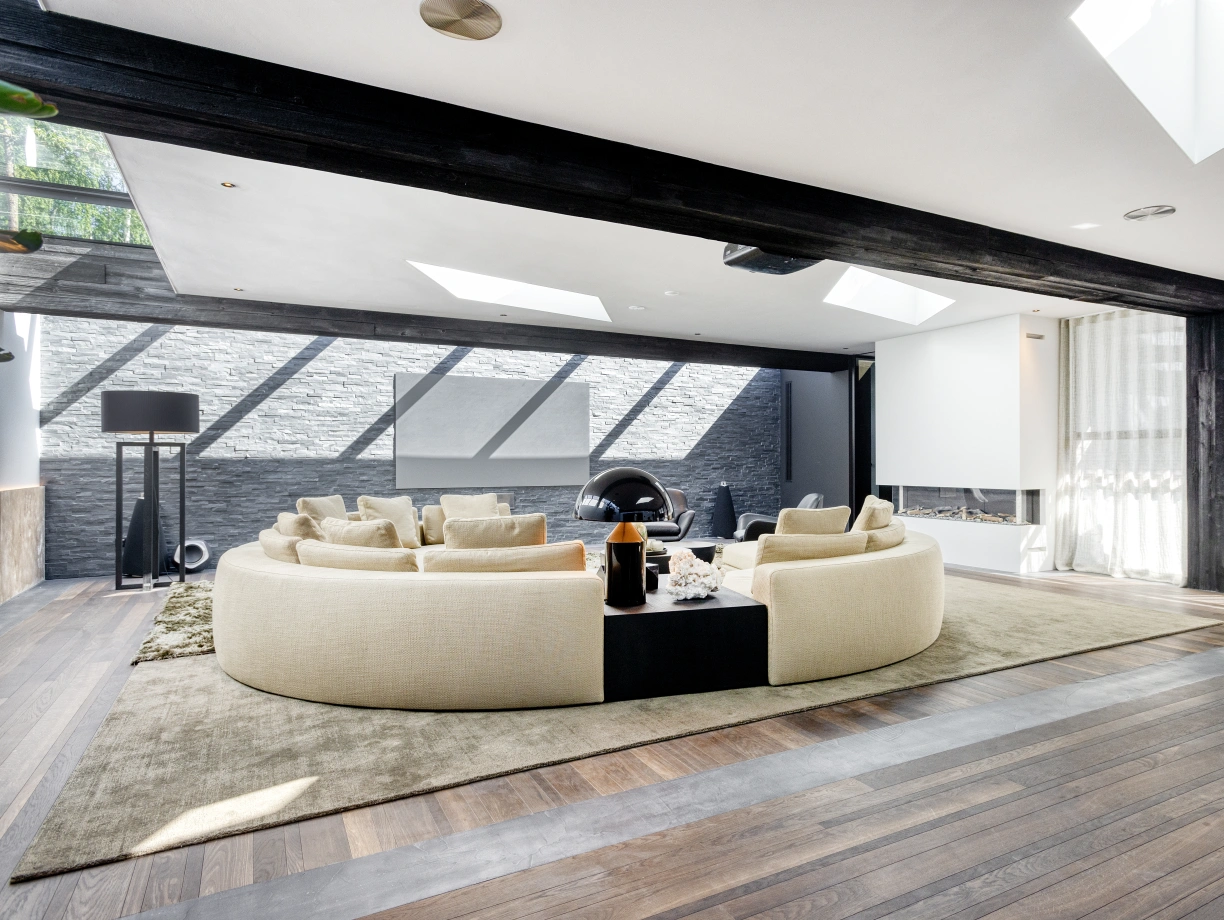 A modern living room featuring a large curved beige sofa arranged around a central coffee table. The space is flooded with natural light from skylights and has a sleek, minimalist design with dark wooden beams and floors. A decorative rug adds warmth, while a cozy reading nook is visible in the background.