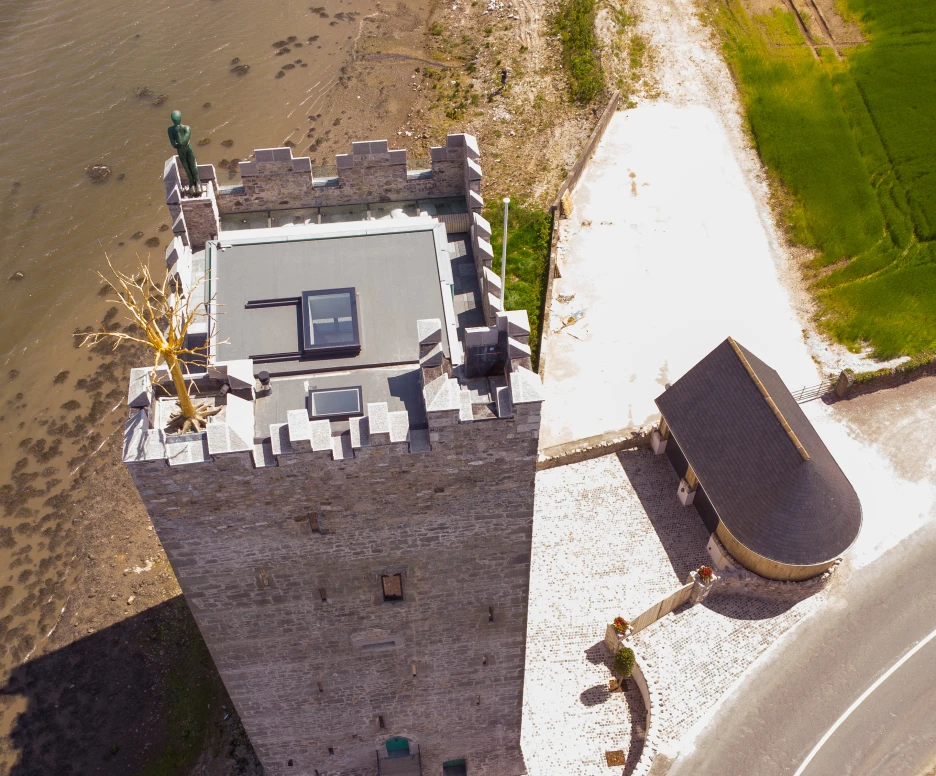 Aerial view of a stone tower with a flat roof and a statue on top, situated near a body of water. A modern building with a curved roof is adjacent to the tower, surrounded by landscaped grounds and a road.
