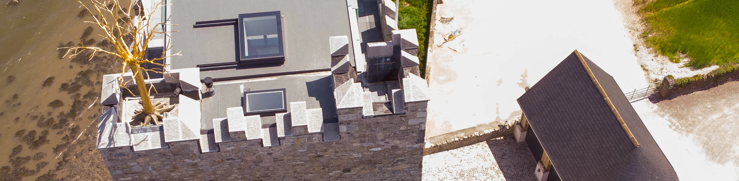 Vista aérea de un castillo de piedra, con un techo plano y una estatua en la parte superior. Al lado, hay una construcción moderna y un camino que rodea la edificación. El entorno es natural, con un área de agua cercana y campos verdes.