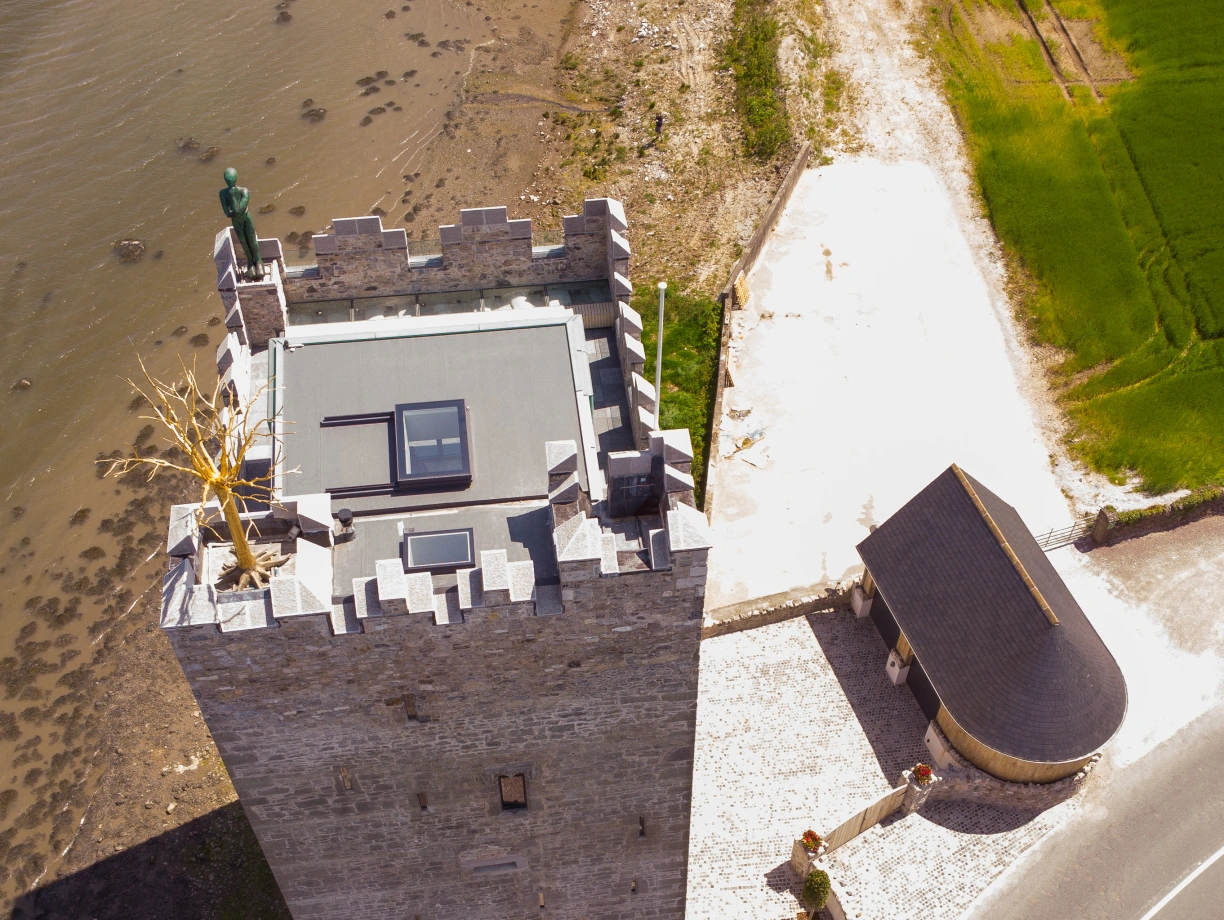 Aerial view of a stone tower with a flat roof, situated near a body of water. A small building with a curved roof is adjacent to the tower. The surrounding area is mostly grass and gravel.