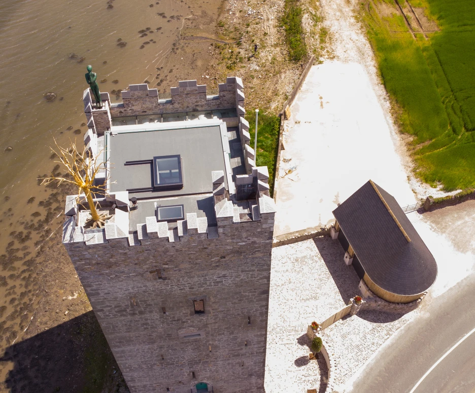 Aerial view of a stone tower with a flat roof, situated near a body of water. A small building with a curved roof is adjacent to the tower. The surrounding area is mostly grass and gravel.