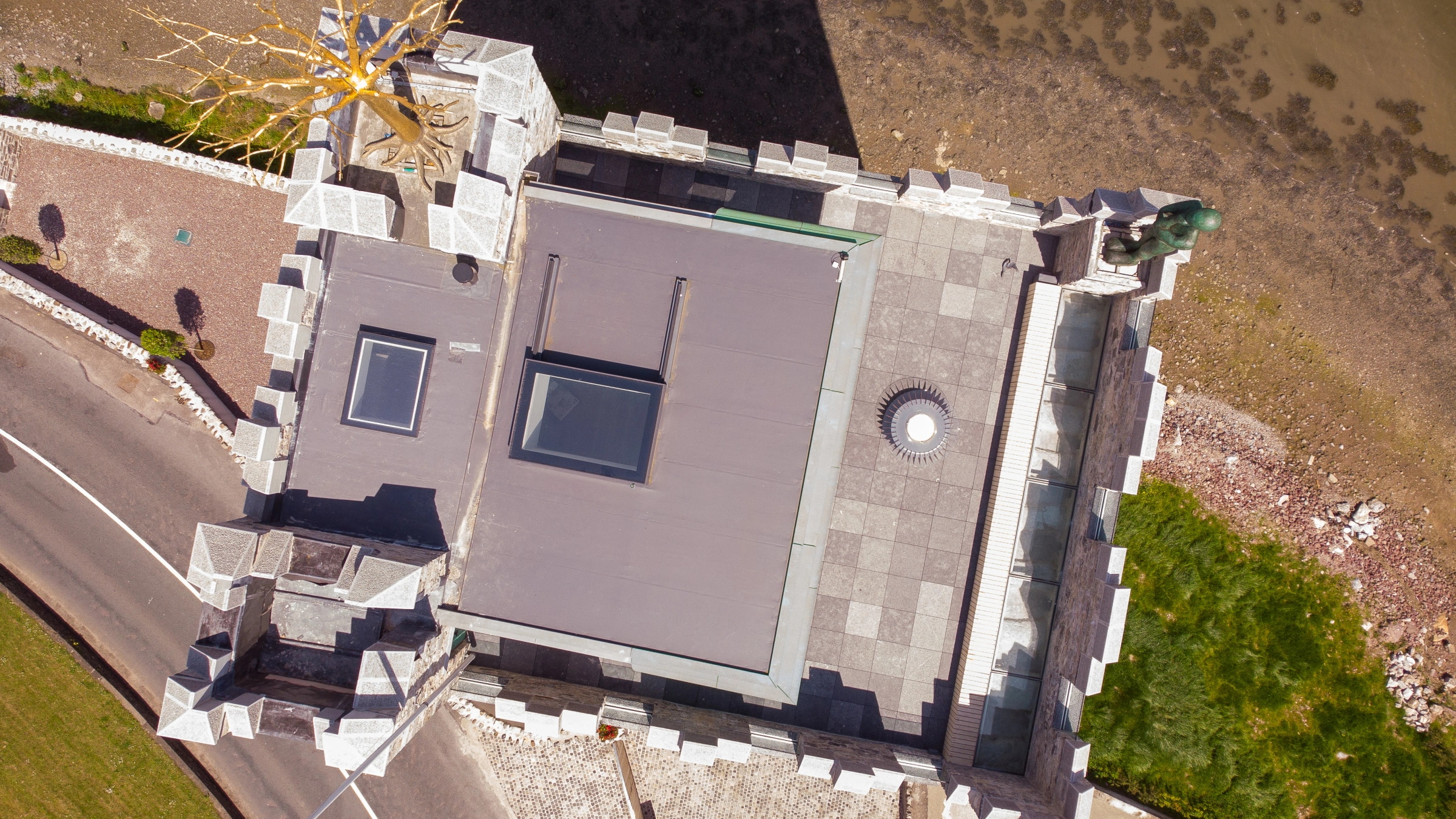 An aerial view of a unique castle-like structure positioned near a shoreline. The roof is flat with skylights, and there is a decorative element resembling a tree. Surrounding the building are stone walls and a road, with a grassy area visible.