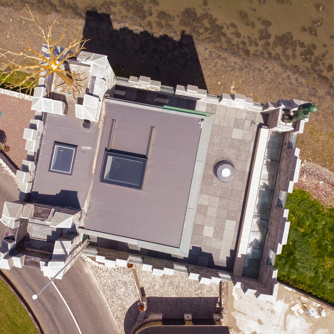 Vista aérea de un castillo con un techo plano y detalles arquitectónicos en la costa. Hay un camino que rodea la estructura y una escultura en una de las esquinas. La vegetación verde contrasta con el entorno costero.