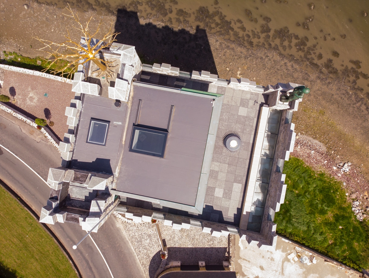 An aerial view of a unique castle-like structure positioned near a shoreline. The roof is flat with skylights, and there is a decorative element resembling a tree. Surrounding the building are stone walls and a road, with a grassy area visible.