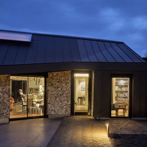 A modern home with a dark metal roof and stone walls is illuminated at dusk. Inside, warm light highlights a dining area and a cozy library. The outdoor space has a simple pathway and soft lighting, enhancing the inviting atmosphere.