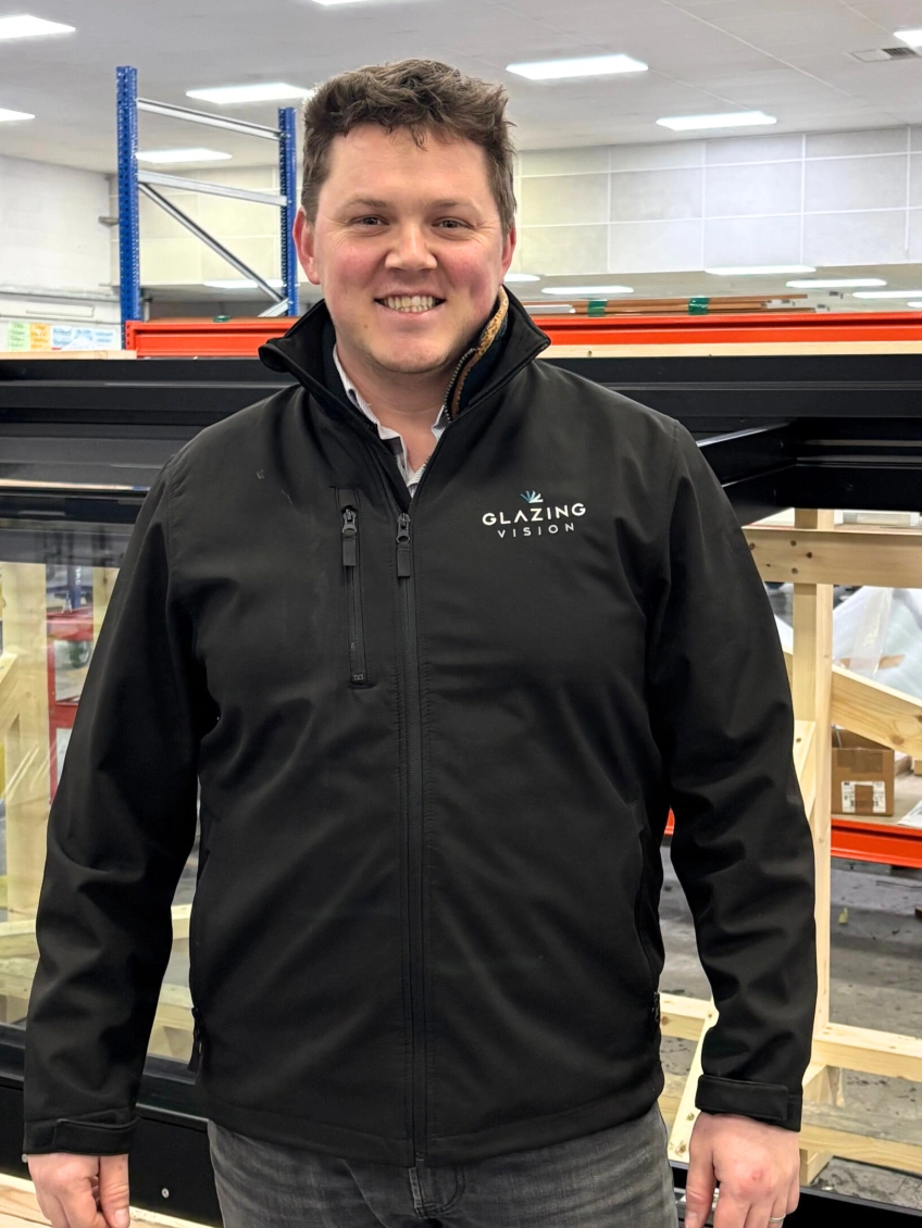 A man wearing a black jacket with the logo "Glazing Vision" stands in a warehouse with wooden structures in the background. He is smiling and looking directly at the camera.