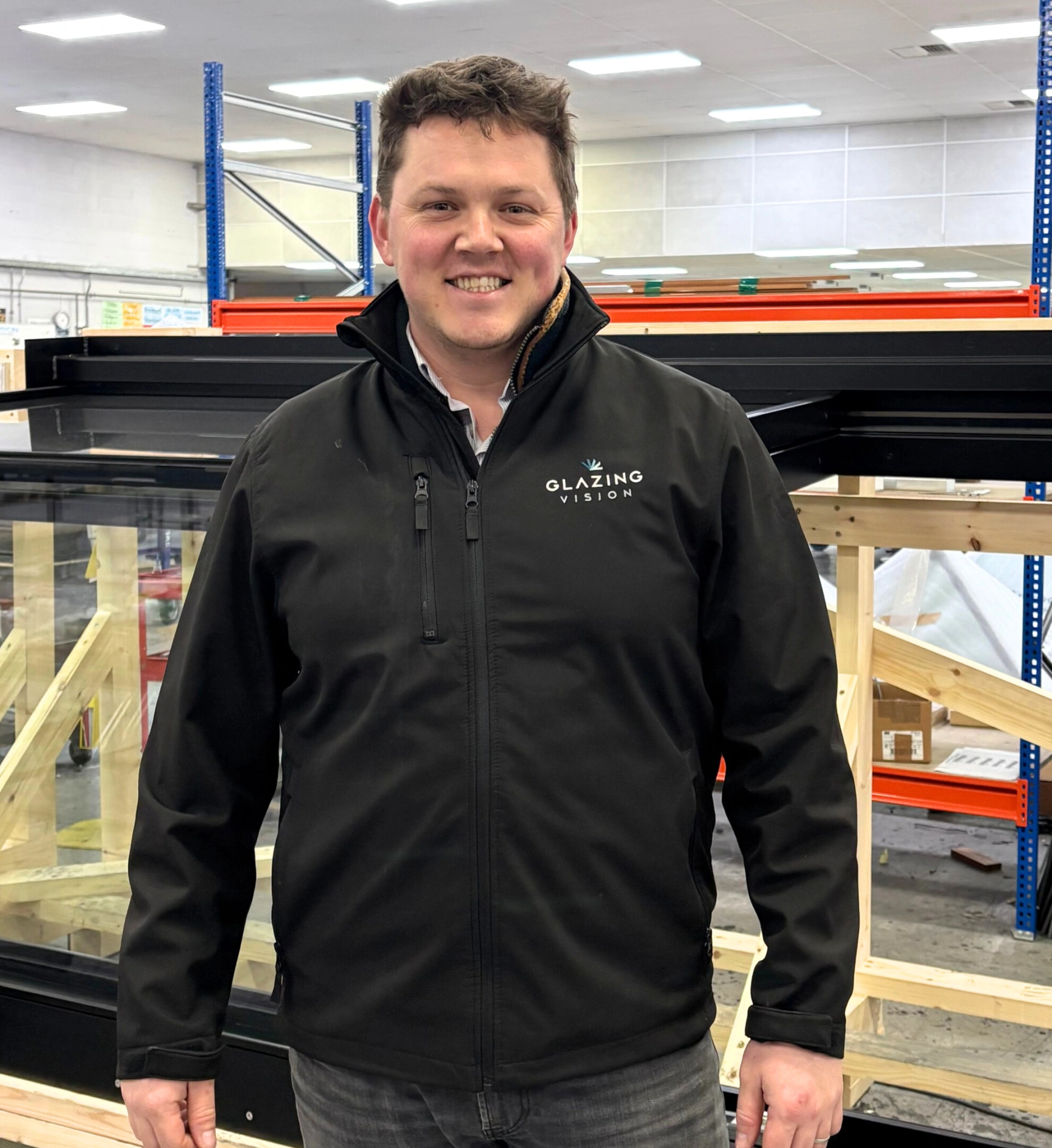 A man wearing a black jacket with the logo "Glazing Vision" stands in a warehouse with wooden structures in the background. He is smiling and looking directly at the camera.