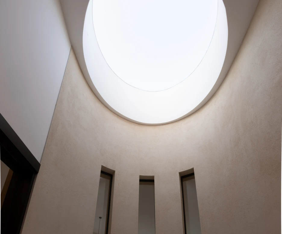 A modern, minimalist interior featuring a circular skylight at the top. Three vertical windows line the walls, allowing natural light to illuminate the space. The walls are smooth and neutral-toned, enhancing the bright atmosphere.