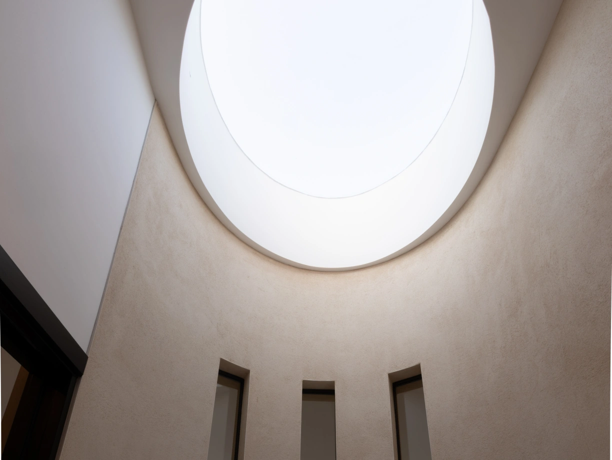 A modern, minimalist interior featuring a circular skylight at the top. Three vertical windows line the walls, allowing natural light to illuminate the space. The walls are smooth and neutral-toned, enhancing the bright atmosphere.