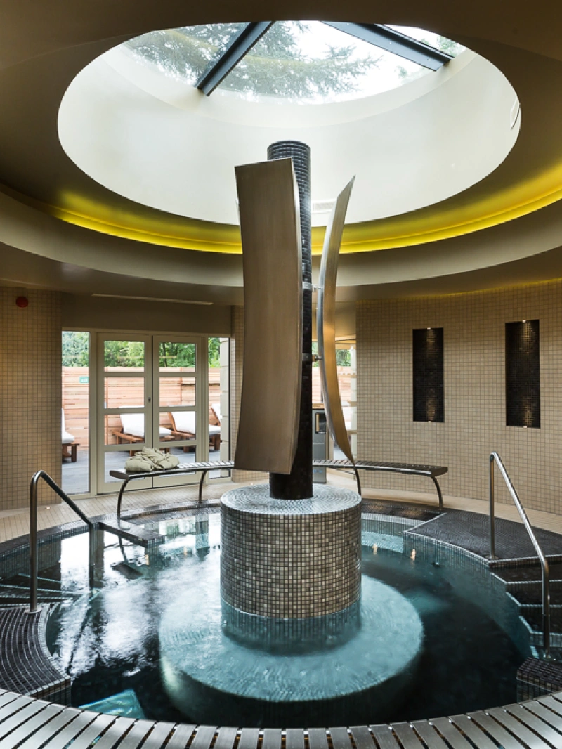 A serene spa interior featuring a circular hot tub with elegant water features, surrounded by mosaic tiles. Natural light streams in through a skylight, illuminating the space, while plush towels are neatly arranged nearby.