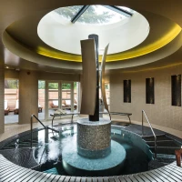 Spa moderno con una fuente central y jacuzzi. La iluminación suave resalta las paredes de azulejos claros y las ventanas permiten la entrada de luz natural. Toallas enrolladas y un ambiente relajante completan la escena.