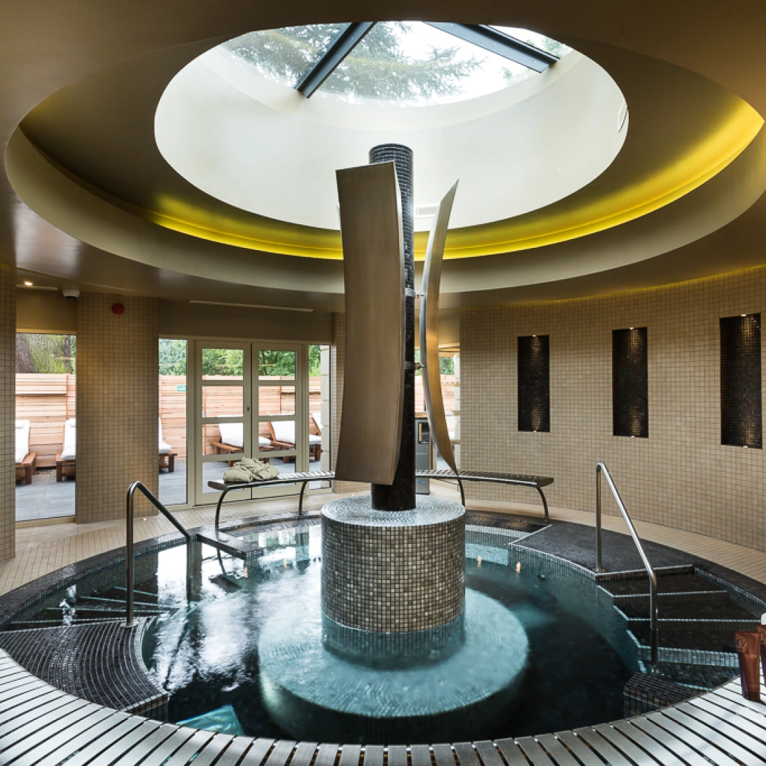 A modern spa interior features a circular skylight and a central water feature with sleek sculptures, surrounded by tiled walls. Comfortable lounge chairs are visible through large glass doors leading outside. Plush towels are neatly arranged on the side.