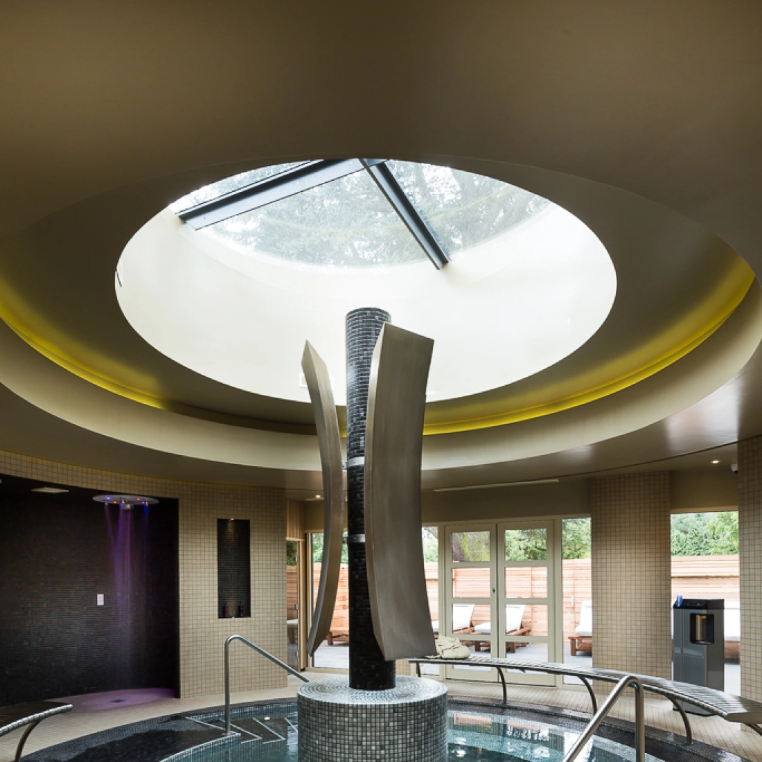 Ein eleganter Spa-Bereich mit einem runden, lichtdurchfluteten Oberlicht. Im Zentrum steht eine moderne Skulptur. Rund um eine Wasserfläche sind Stufen aus dunklen Fliesen und entspannende Liegen angeordnet.