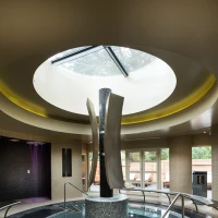 Ein eleganter Spa-Bereich mit einem runden, lichtdurchfluteten Oberlicht. Im Zentrum steht eine moderne Skulptur. Rund um eine Wasserfläche sind Stufen aus dunklen Fliesen und entspannende Liegen angeordnet.