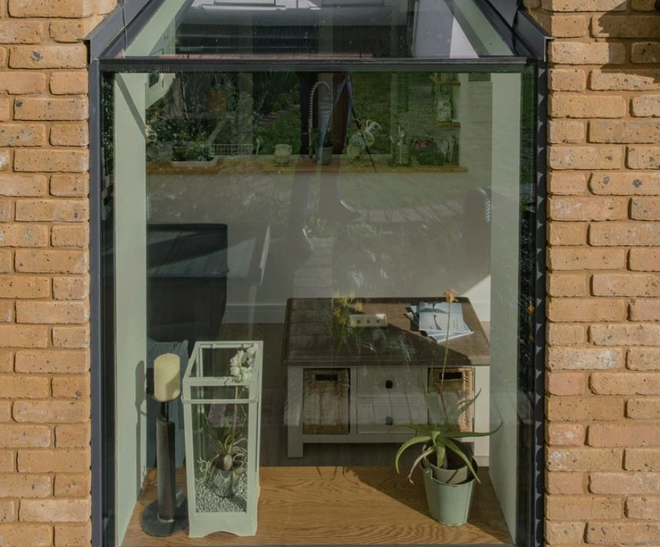 A modern glass extension with a sloped roof, showcasing wooden flooring, a small coffee table, and a green plant. The walls are made of light-colored brick, creating a cozy atmosphere with natural light.