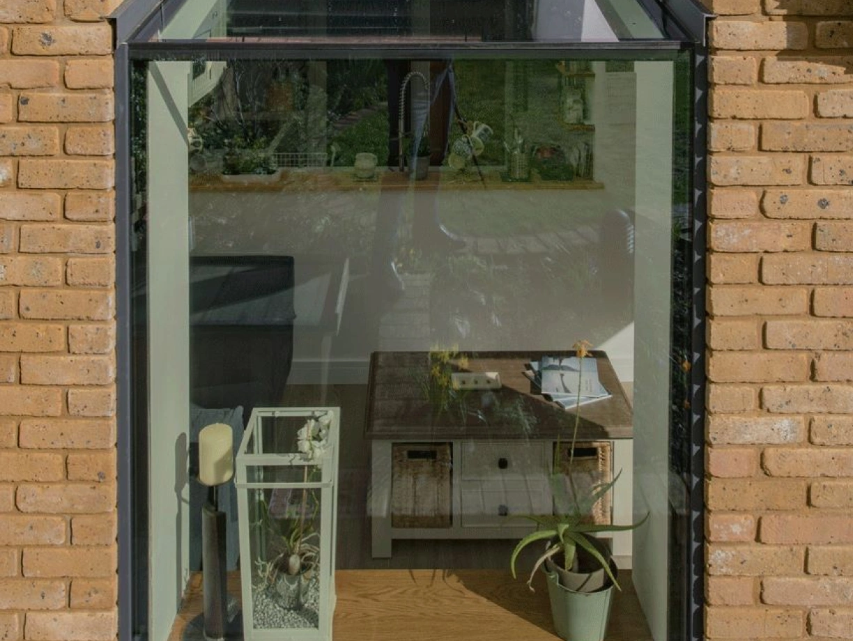 A modern glass extension with a sloped roof, showcasing wooden flooring, a small coffee table, and a green plant. The walls are made of light-colored brick, creating a cozy atmosphere with natural light.