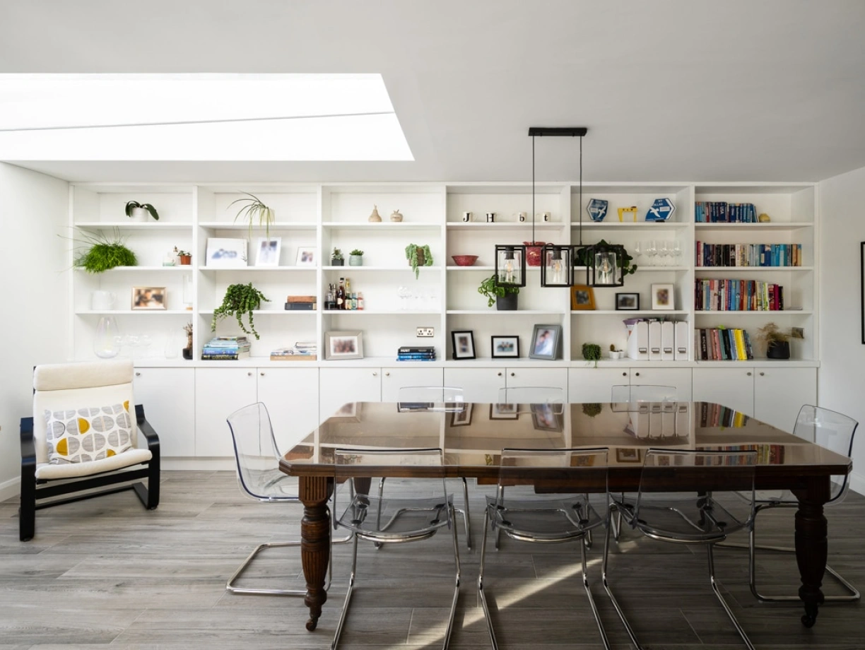 A modern dining area featuring a large wooden table surrounded by transparent chairs. Behind the table, there is a white shelving unit filled with books, plants, and framed photos. Natural light streams in from a skylight above.
