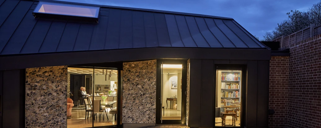 A modern home exterior at dusk, featuring a unique angular roof and large glass doors. Inside, glimpses of a cozy dining area and a bookshelf are visible, illuminated warmly. The surrounding landscape is serene, with a pathway leading to a softly lit outdoor area.