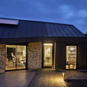 A modern home exterior at dusk, featuring a unique angular roof and large glass doors. Inside, glimpses of a cozy dining area and a bookshelf are visible, illuminated warmly. The surrounding landscape is serene, with a pathway leading to a softly lit outdoor area.