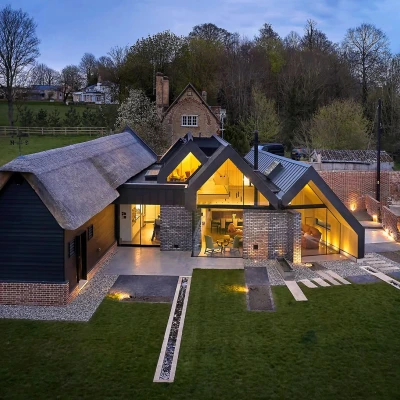A modern house featuring a combination of thatched and metal roofs, surrounded by a landscaped yard with pathways. The structure has large windows that illuminate the interior at dusk. Countryside trees and distant buildings are visible in the background.