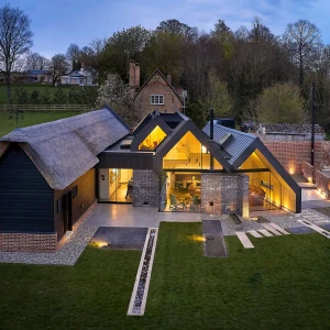 A modern house featuring a combination of thatched and metal roofs, surrounded by a landscaped yard with pathways. The structure has large windows that illuminate the interior at dusk. Countryside trees and distant buildings are visible in the background.