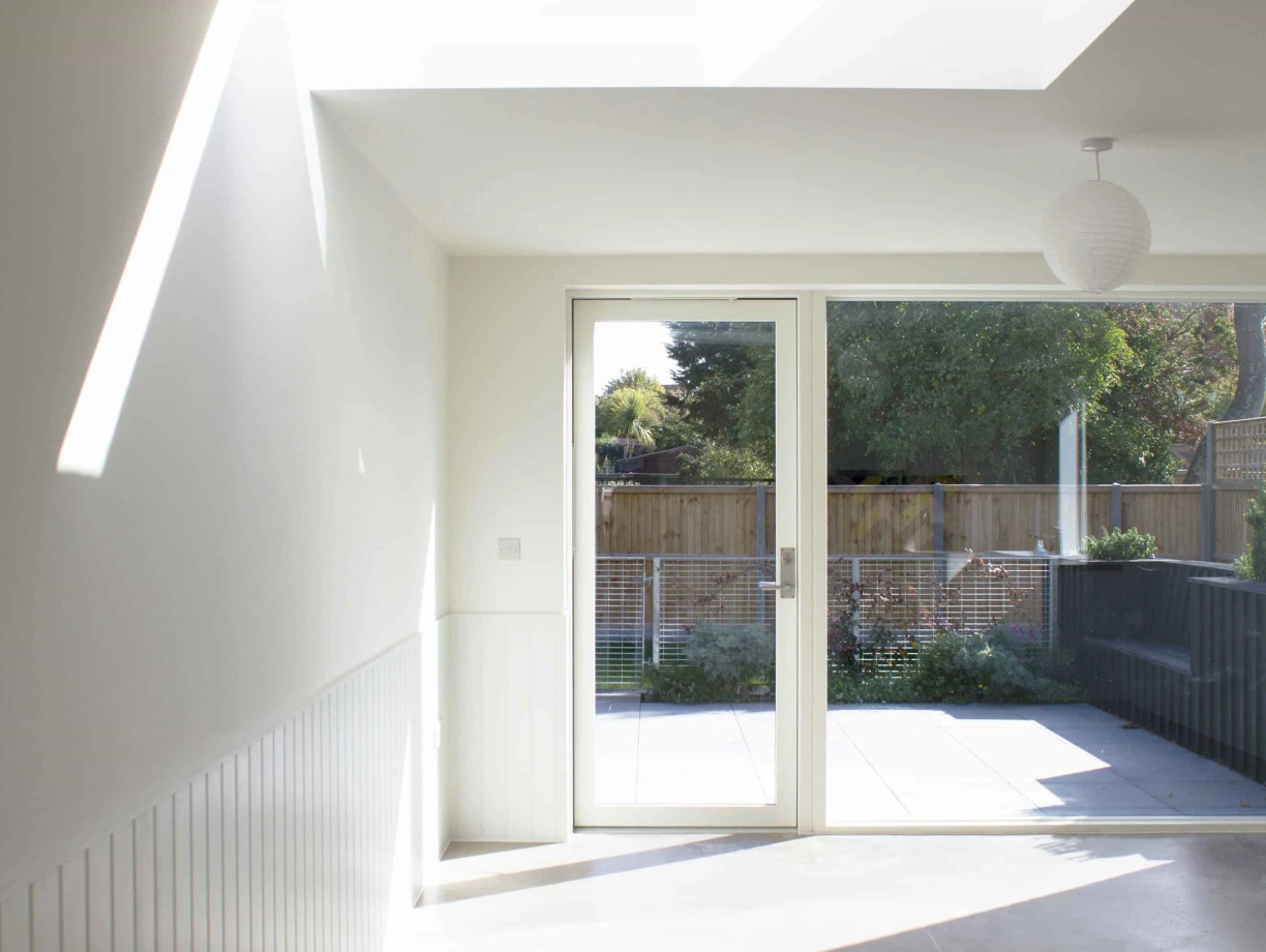 Bright, modern interior with a large glass door leading to an outdoor patio. Large skylight allows natural light to fill the space. Minimalist design features white walls and a subtle pendant light. Greenery is visible outside the door.