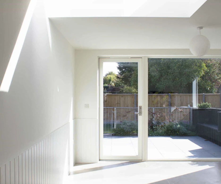 Bright, modern interior with a large glass door leading to an outdoor patio. Large skylight allows natural light to fill the space. Minimalist design features white walls and a subtle pendant light. Greenery is visible outside the door.