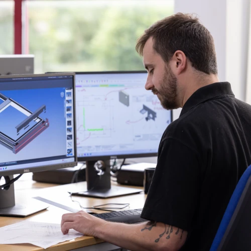A man is seated at a desk, working on computer-aided design (CAD) software on dual monitors. He is reviewing technical drawings and appears focused. The office has modern furnishings and natural light.