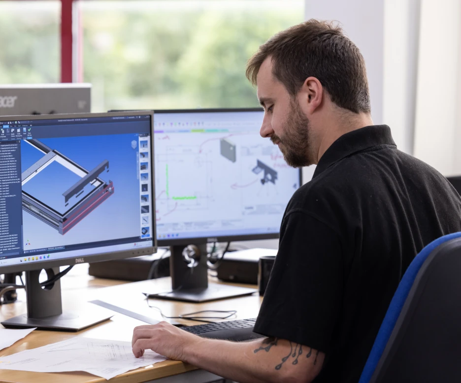 A man sitting at a desk, focusing on a computer screen displaying a 3D model and engineering designs. He has a notepad and documents in front of him. Two additional monitors are visible in the background.