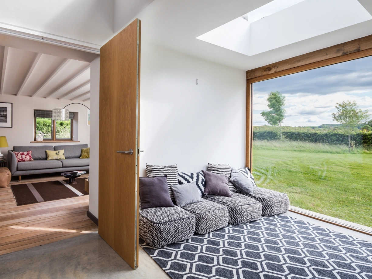 A bright and modern interior featuring a cozy seating area with patterned cushions and a geometric rug. A wooden door opens to a spacious room with a sofa, decorative lamp, and framed artwork. Large windows provide a view of a green landscape and a blue sky.