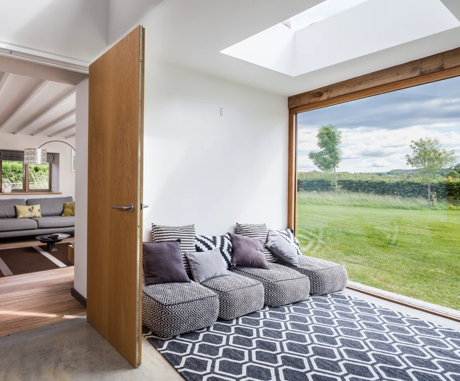 A bright and modern interior featuring a cozy seating area with patterned cushions and a geometric rug. A wooden door opens to a spacious room with a sofa, decorative lamp, and framed artwork. Large windows provide a view of a green landscape and a blue sky.
