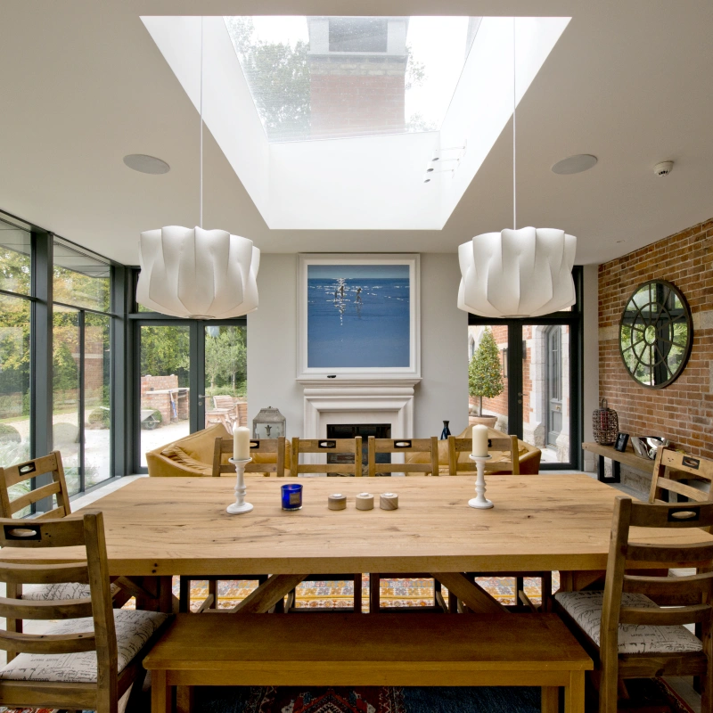 A modern dining room features a large wooden table surrounded by chairs. Two stylish white pendant lights hang above the table, and a large skylight allows natural light to fill the space. A fireplace with a simple design contrasts with the brick wall. Large glass doors offer a view of the outdoor area.