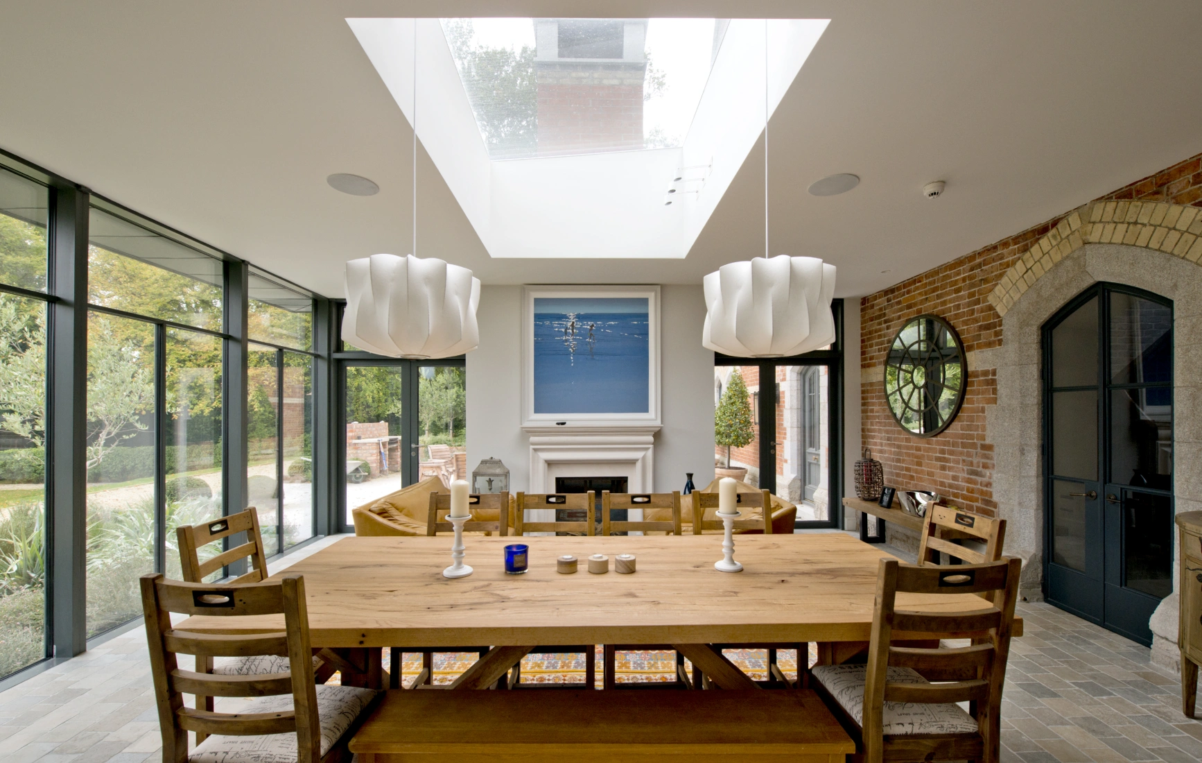 A modern dining room features a large wooden table surrounded by chairs. Two stylish white pendant lights hang above the table, and a large skylight allows natural light to fill the space. A fireplace with a simple design contrasts with the brick wall. Large glass doors offer a view of the outdoor area.