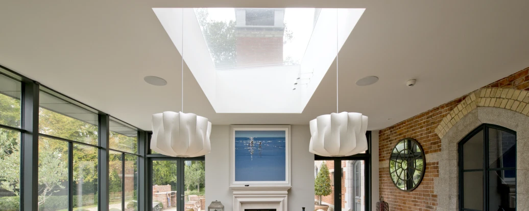 Bright, modern dining room featuring a large wooden table with eight chairs, soft lighting from two unique pendant fixtures, and a central artwork above a fireplace. Large glass windows offer views of the garden. A skylight brings in natural light, enhancing the open and airy atmosphere.