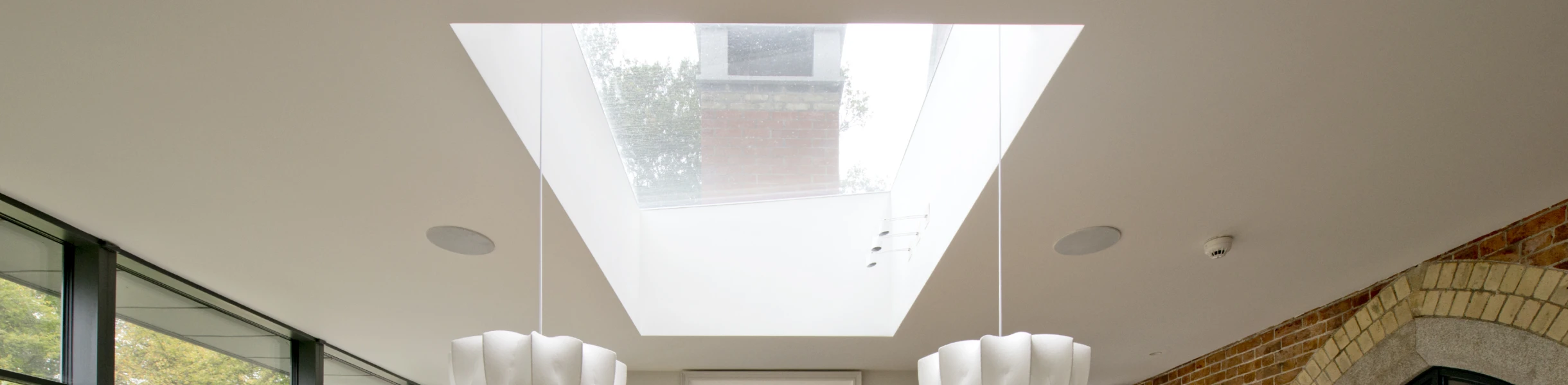 Ein moderner Essbereich mit einem großen Holztisch und Stühlen. Über dem Tisch hängen stilvolle Lampen. An einer Wand befindet sich ein großes Fenster und ein Kamin. Im Hintergrund sind Pflanzen und ein Ausblick auf einen Garten sichtbar.