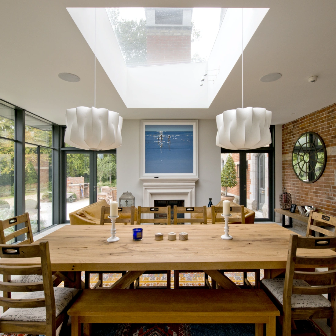 Bright, modern dining room featuring a large wooden table with eight chairs, soft lighting from two unique pendant fixtures, and a central artwork above a fireplace. Large glass windows offer views of the garden. A skylight brings in natural light, enhancing the open and airy atmosphere.