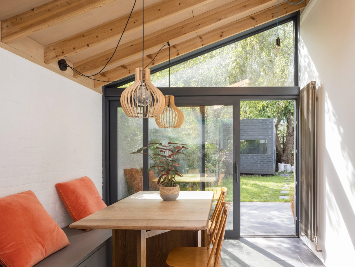 A modern dining area featuring a wooden table surrounded by chairs, with orange cushions on a bench. Large glass doors open to a green outdoor space, illuminated by natural light from a slanted wooden ceiling adorned with stylish hanging lights.