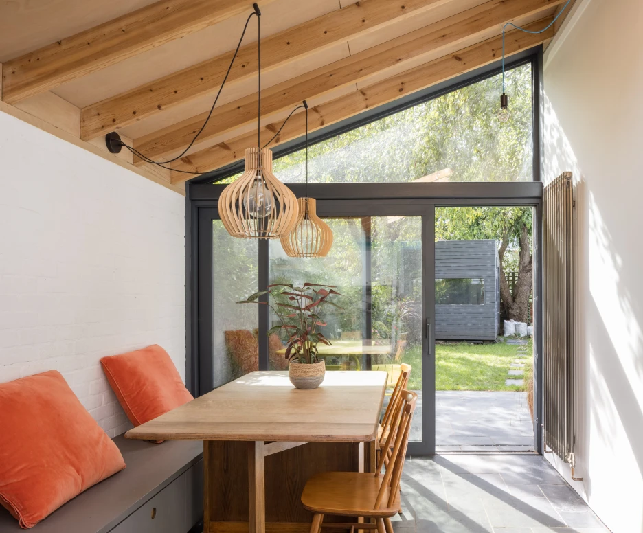 A modern dining area featuring a wooden table surrounded by chairs, with orange cushions on a bench. Large glass doors open to a green outdoor space, illuminated by natural light from a slanted wooden ceiling adorned with stylish hanging lights.