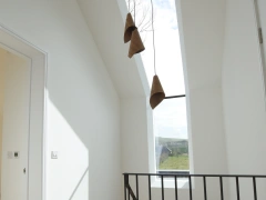 A bright hallway with a modern design, featuring a large skylight and three pendant lights hanging from the ceiling. There is a black railing along the staircase leading down, and the walls are painted white, creating a clean and airy atmosphere.