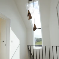 Un couloir lumineux avec un plafond en pente et une grande fenêtre ouverte sur le ciel. Trois suspensions en forme de cône sont accrochées au plafond. Un escalier en bois est visible à gauche, avec une balustrade noire minimaliste.