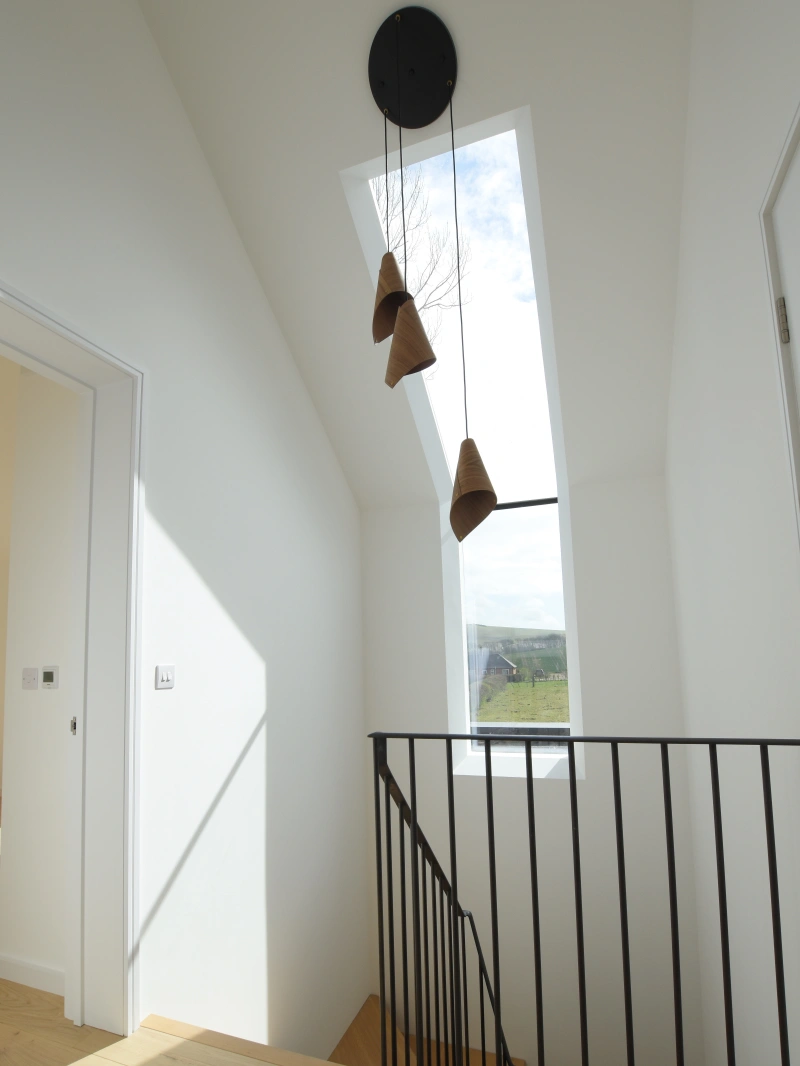 A modern hallway with a sloped ceiling features three pendant lights hanging from a skylight. A black railing borders the wooden staircase, and sunlight streams through the window, casting shadows on the floor.