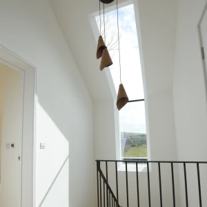 A bright hallway with a modern design, featuring a large skylight and three pendant lights hanging from the ceiling. There is a black railing along the staircase leading down, and the walls are painted white, creating a clean and airy atmosphere.