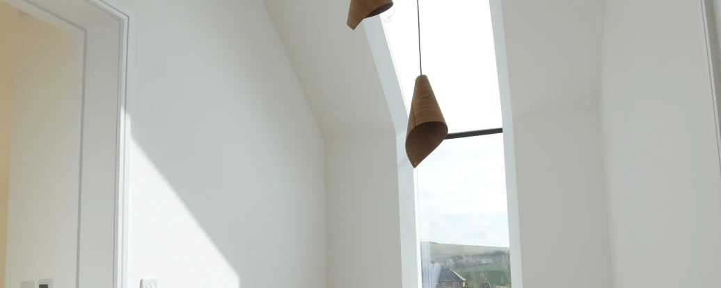 A bright hallway with a modern design, featuring a large skylight and three pendant lights hanging from the ceiling. There is a black railing along the staircase leading down, and the walls are painted white, creating a clean and airy atmosphere.