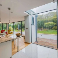Modern kitchen area featuring a granite countertop, a vase of flowers, and sleek pendant lighting. In the background, a cozy living space with colorful furniture opens up to a lush garden through large glass doors. Bright and airy atmosphere with natural light.