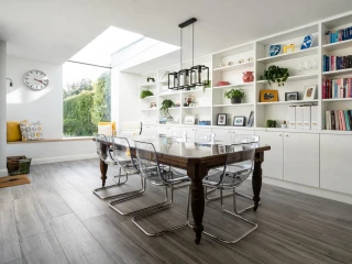 A modern dining area features a wooden table surrounded by clear acrylic chairs. Shelves filled with books, plants, and decorative items line one wall. A skylight brings in natural light, highlighting the spacious, airy atmosphere. A cozy nook with cushions is visible in the background.