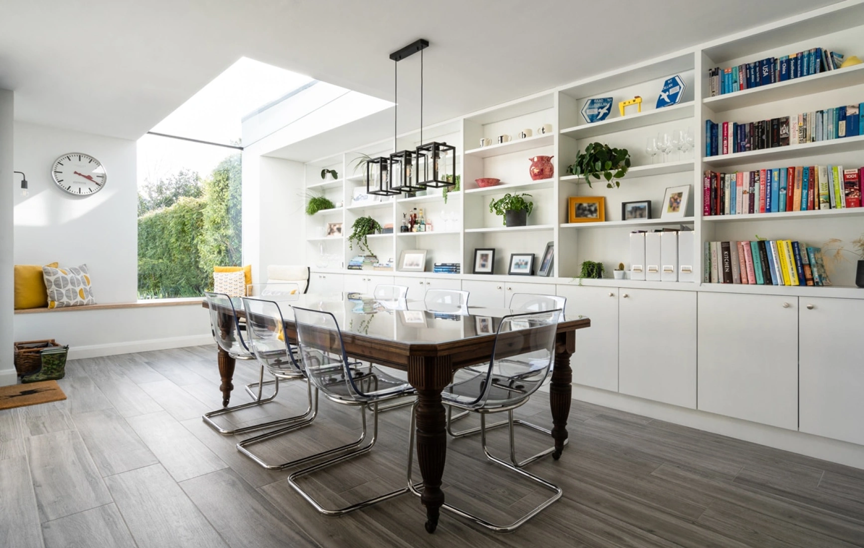 In a dining area with a table in the centre with glass chairs all around it. To the right there is a long whiite book shelf going across the whole wall which is filled with books and plants. To the left there is a sitting area with yellow cushions and a big clock above which is next to an eaves skylight.