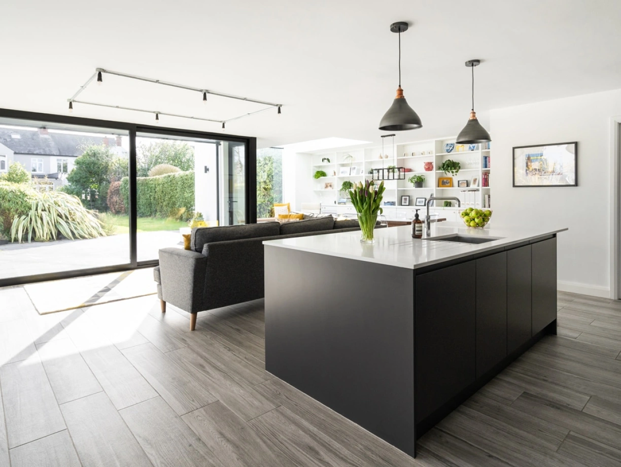 A modern kitchen featuring a large island with a white countertop, dark cabinetry, and pendant lights. There is a comfortable seating area with a gray sofa in the background, lush plants, and sliding glass doors leading to a garden. Brightly colored shelves display decorative items.